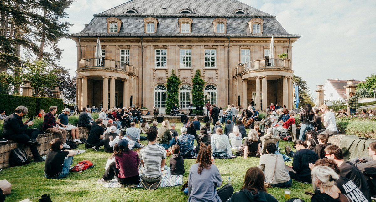 Menschen sitzen auf dem Rasen vor der Villa Reitzenstein und hören einer Person zu.